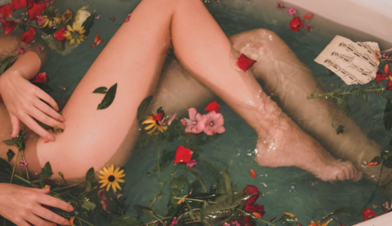 white bathtub filled with water. white female arms with crooked legs. the water is filled with pink, yellow and red flowers and paper
