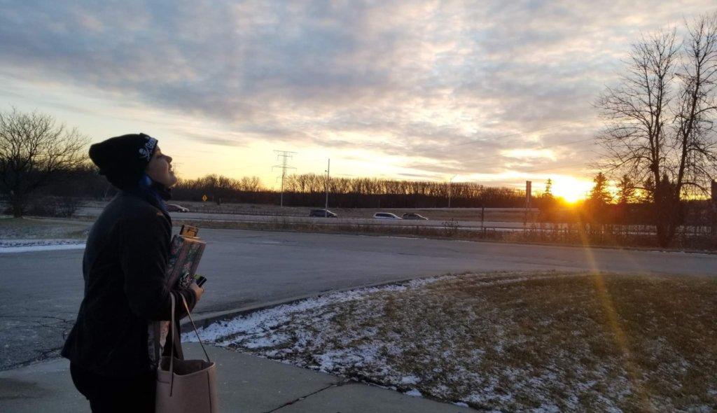 a late afternoon sky with white clouds and a setting sun and trees in the distance. a young woman in a beanie holding books and a bag is throwing her head back and looking towards the sky